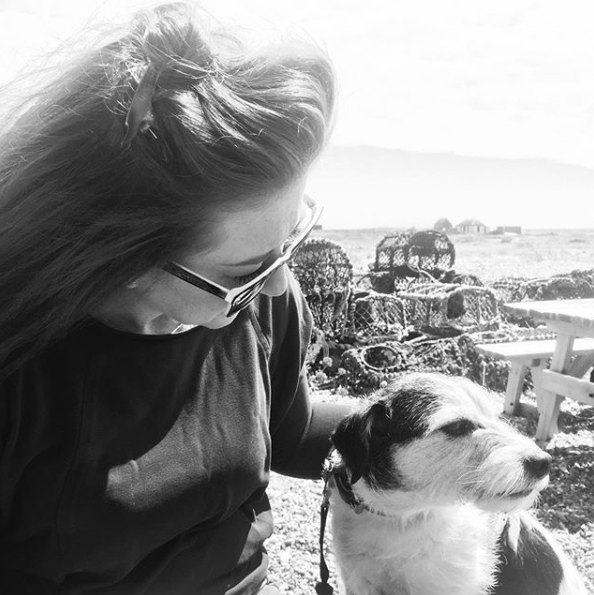 Susanne Wakefield Susanne is wearing sunglasses and has her head tilted down to the right looking at a dog. The dog is mostly white with patches of darker, possibly black or dark brown coat. They are outside. Behind them there is a picnic table and a pile of lobster pots, indicating they are at the coast.